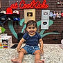 toddler, child, smiling, countertop, brick_wall, neon_sign, graffiti, play_buttons, hats, plush_toys, store_interior, shorts, tshirt, bracelets, footwear_store, colorful, indoor, person, happy, cute