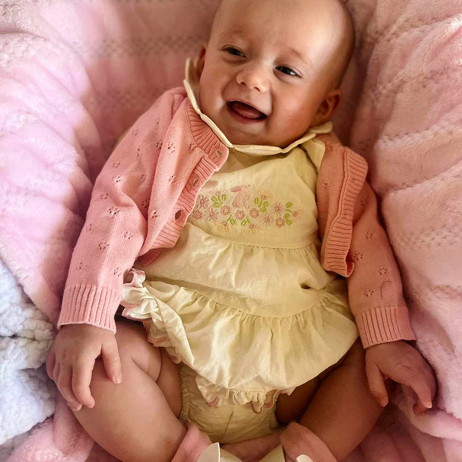 Layla joined the competition — help win amazing prizes! baby, baby_shoes, blanket, child, comfortable, cozy, cream_dress, cute, happy, indoors, infant, person, pink_cardigan, portrait, sitting, smiling, soft_texture, toddler, warm_colors, young_child