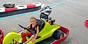Bradley participe au concours pour gagner de l'argent avec cette photo : asphalt, barrier, black, boy, child, fence, go_kart, green, happy, helmet, outdoor, race_track, racing, recreation, red, shorts, smile, sportswear, vehicle, yellow