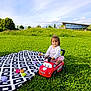 blanket, casual_clothing, child, clouds, daytime, fun, grass, greenery, happy, nature, outdoor, park, play, sky, smiling, sunlight, toddler, toy, toy_car, young_child