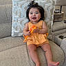 Naomi is registered to the contest to win money with this photo: baby, child, smile, happy, toy, orange, yellow, couch, cushion, indoor, home, furniture, person, hair, clothing, sitting, cute, infant, playful, relaxed