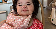 Naomi joined the competition — help win amazing prizes! baby, high_chair, bib, teething_toy, food, messy_face, red_checkered_clothing, pink_floral_bib, indoor, kitchen, living_room, child, person, smiling, cute, hair, hand, table, furniture, home