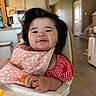 baby, high_chair, bib, teething_toy, food, messy_face, red_checkered_clothing, pink_floral_bib, indoor, kitchen, living_room, child, person, smiling, cute, hair, hand, table, furniture, home