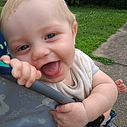 Eric is registered to the contest to win money with this photo: baby, child, smiling, tongue_out, stroller, hand, face, blue_eyes, outdoor, grass, sidewalk, happy, cute, infant, person, clothing, playful, skin, head, nature