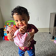 Elizabeth is registered to the contest to win money with this photo: toddler, child, baby_doll, smiling, curly_hair, indoor, carpet, barefoot, toy, furniture, playtime, happy, person, holding, cute, young_child, clothing, living_room, cabinet, floor
