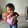 toddler, child, baby_doll, smiling, curly_hair, indoor, carpet, barefoot, toy, furniture, playtime, happy, person, holding, cute, young_child, clothing, living_room, cabinet, floor