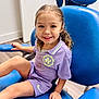 child, smile, purple_clothing, necklace, curly_hair, blue_chair, pigtails, indoor, wood_floor, door, person, happy, sitting, young_girl, casual_wear, bright, clean, room, portrait, cute