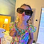 child, girl, sunglasses, floral_dress, bedroom, lamp, bed, puckered_lips, curly_hair, indoor, playful, cute, fashion, portrait, childhood, smile, person, home, light, furniture