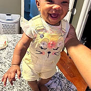 Leilani joined the competition — help win amazing prizes! baby, child, smiling, yellow_overalls, bed, hand, indoor, person, happy, clothing, foot, face, cute, infant, home, pattern, fabric, floor, skin, holding