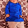 baby, child, blue_clothing, traditional_outfit, bedspread, patterned_fabric, indoor, lying_down, footwear, curious_expression, face, hands, pajamas, brown_pattern, white_pants, young_child, portrait, cute, indoors, resting