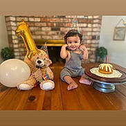 Iker joined the competition — help win amazing prizes! baby, balloon, birthday, birthday_cake, brick_wall, cake_stand, candle, celebration, child, fireplace, gold_balloon, indoor, party_hat, portrait, sitting, smiling, table, teddy_bear, text_overlay, wooden_table