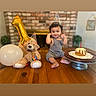 baby, balloon, birthday, birthday_cake, brick_wall, cake_stand, candle, celebration, child, fireplace, gold_balloon, indoor, party_hat, portrait, sitting, smiling, table, teddy_bear, text_overlay, wooden_table