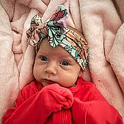 Emberleigh is registered to the contest to win money with this photo: baby, infant, headband, red_clothing, blanket, soft_texture, cute, portrait, child, newborn, face, hands, lying_down, colorful, floral_pattern, cozy, warm, pink, closeup, expression