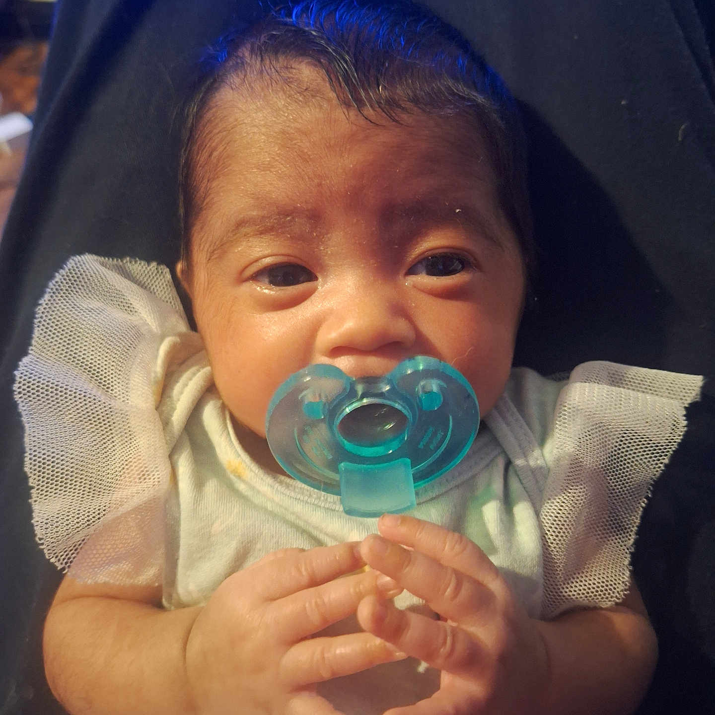 Vivian is registered to the contest to win money with this photo: baby, pacifier, infant, hands, clothing, frilly_sleeves, lap, person, dark_pants, closeup, face, curious, skin, hair, light, indoors, cute, newborn, resting, portrait