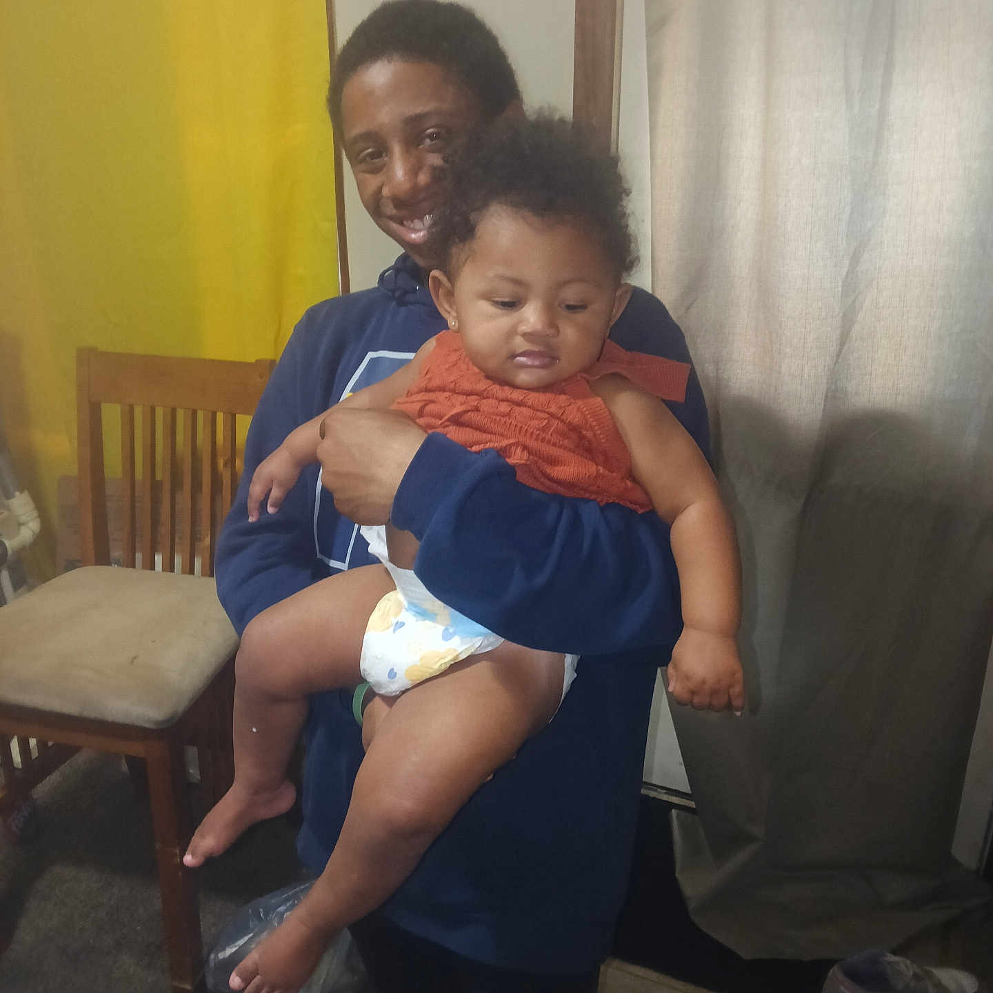 Alina is registered to the contest to win money with this photo: adult, baby, child, diaper, orange_clothing, holding, smiling, indoor, curtain, chair, carpet, socks, shoes, person, family, happy, toddler, furniture, wall, portrait