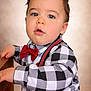 toddler, child, boy, face, blue_eyes, curly_hair, red_bow_tie, suspenders, checkered_shirt, long_sleeve, portrait, indoor, wooden_surface, curious_expression, close_up, fashion, cute, young_child, casual_clothing, soft_background
