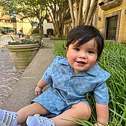 Robert is registered to the contest to win money with this photo: toddler, child, boy, smiling, blue_clothing, white_sneakers, grass, sidewalk, urban, trees, potted_plant, brick_building, outdoor, daylight, face, happy, sitting, cute, short_hair, casual