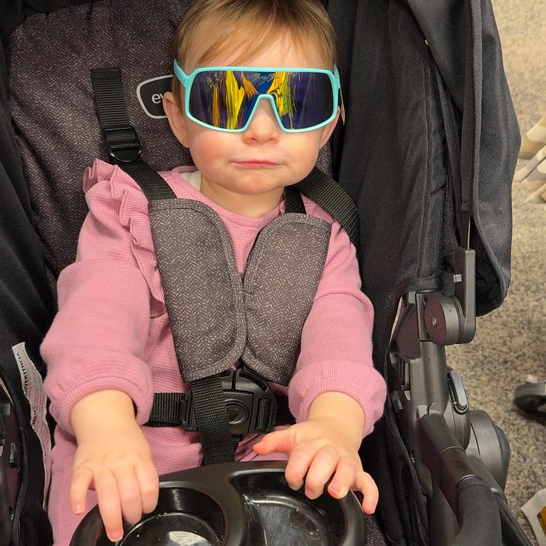 Baylin is registered to the contest to win money with this photo: toddler, child, sunglasses, stroller, pink_clothing, baby, seatbelt, indoor, person, footwear, tray, hand, face, head, expression, toy, casual, cute, fashion, transport