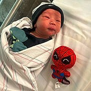 Miles joined the competition — help win amazing prizes! newborn, baby, hospital, blanket, beanie, plush_toy, spiderman, bed, infant, cute, wrapped, child, soft_toy, lying_down, person, black_clothing, white_bed, baby_mittens, portrait, indoors