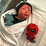 newborn, baby, hospital, blanket, beanie, plush_toy, spiderman, bed, infant, cute, wrapped, child, soft_toy, lying_down, person, black_clothing, white_bed, baby_mittens, portrait, indoors
