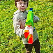 Alfie is registered to the contest to win money with this photo: child, toy_gun, water_gun, green_grass, outdoor, playful, sunny, hoodie, casual_clothing, red_shoes, fence, garden, daylight, young_boy, grass, greenery, person, smiling, fun, nature