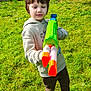 child, toy_gun, water_gun, green_grass, outdoor, playful, sunny, hoodie, casual_clothing, red_shoes, fence, garden, daylight, young_boy, grass, greenery, person, smiling, fun, nature