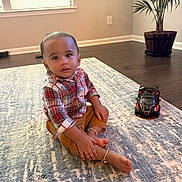 Vihansh is registered to the contest to win money with this photo: toddler, child, indoor, carpet, toy_car, plaid_shirt, pants, barefoot, bracelets, floor, plant, window, light, living_room, person, furniture, wood_floor, curious, sitting, home