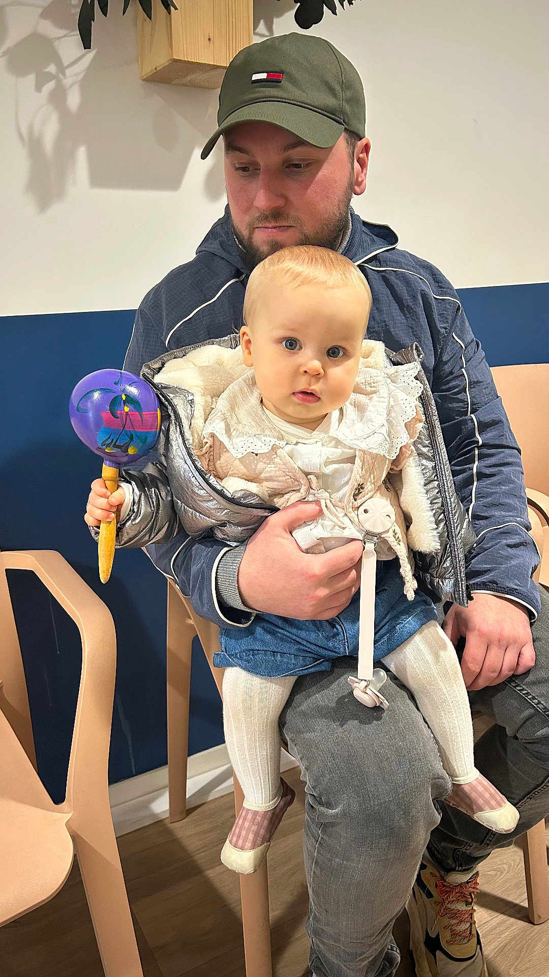 Juna a rejoint le concours — aidez-le/la à gagner de superbes lots ! baby, man, child, indoor, seated, rattle, jacket, cap, toy, plastic_chair, floor, person, clothing, footwear, wall, wood, sitting, expression, holding, curious