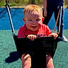 toddler, child, swing, playground, happy, smiling, outdoor, sunny, red_hair, black_swing, grass, adult, blurred_background, holding, seated, play, fun, daylight, casual_clothing, park