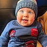 baby, child, blue_eyes, knitted_hat, red_pom_pom, spiderman_outfit, sweater, pants, smiling, cute, indoor, seated, blanket, mustard_color, soft_lighting, face, headwear, winter_clothing, portrait, infant