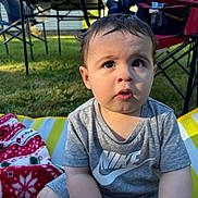 Rey is registered to the contest to win money with this photo: toddler, child, outdoor, grass, blanket, striped_mat, nike_shirt, curious_expression, folding_chair, sunlight, short_hair, sitting, person, daytime, casual_clothing, greenery, summer, face, eyes, mouth