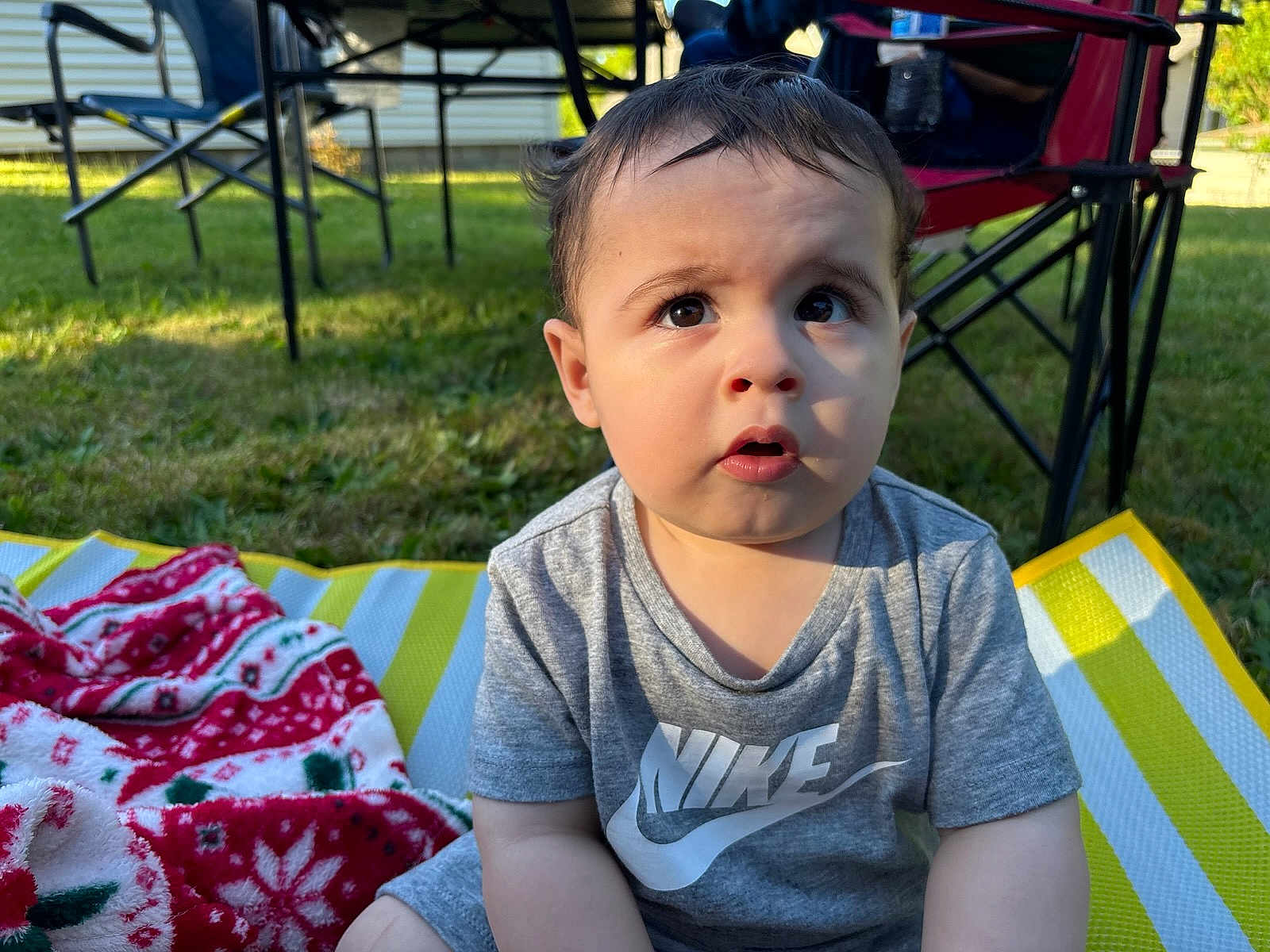 Rey is registered to the contest to win money with this photo: toddler, child, outdoor, grass, blanket, striped_mat, nike_shirt, curious_expression, folding_chair, sunlight, short_hair, sitting, person, daytime, casual_clothing, greenery, summer, face, eyes, mouth