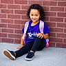 child, sitting, brick_wall, purple_shirt, pink_backpack, curly_hair, smile, black_shoes, black_leggings, outdoor, concrete_floor, casual_clothing, young_girl, portrait, daylight, happy, resting, fashion, person, cute