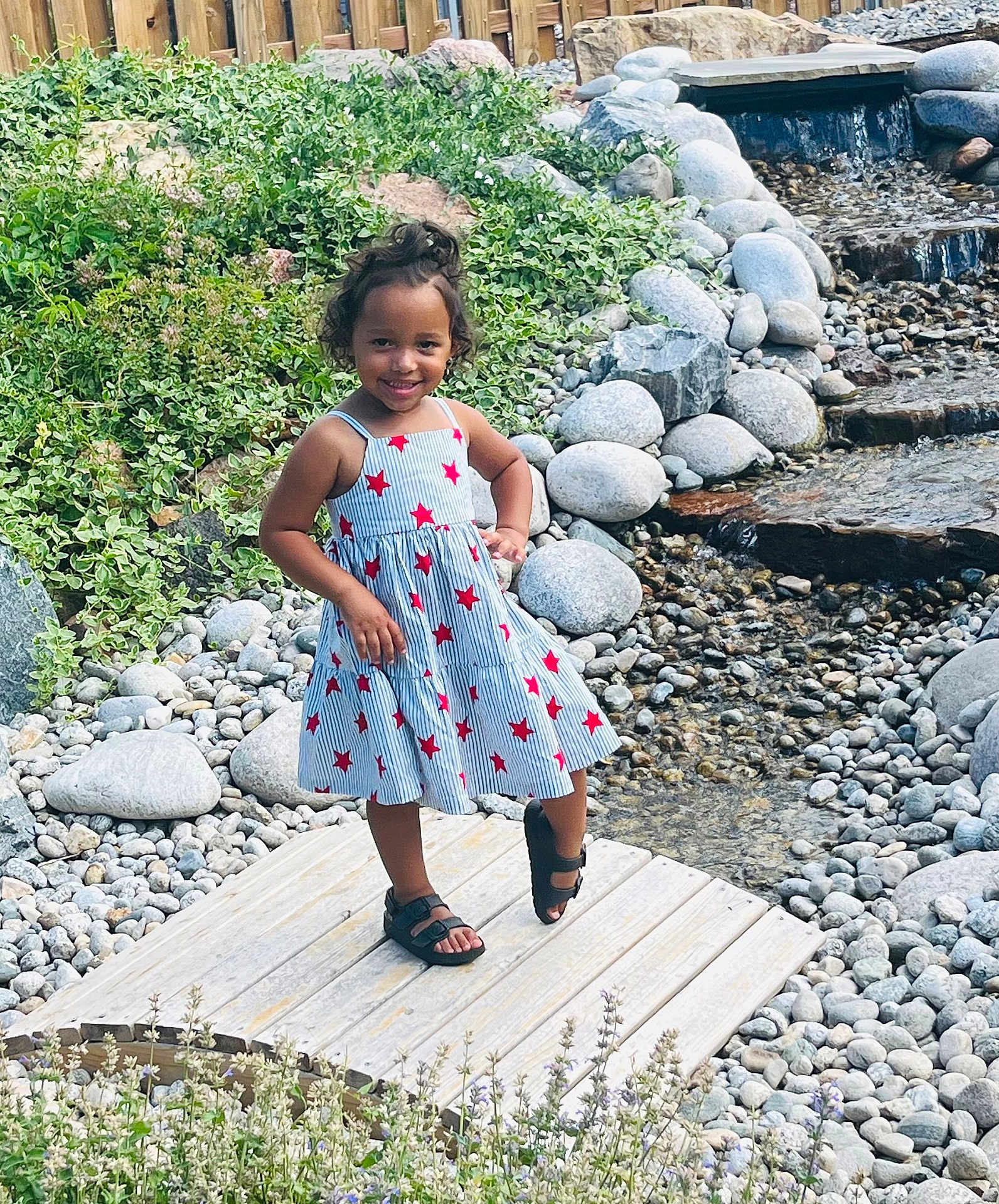Zai'Lynn joined the competition — help win amazing prizes! child, girl, smiling, dress, stars, sandals, outdoor, garden, rocks, water, stream, plants, wooden_platform, nature, happy, posing, sunlight, greenery, young, portrait
