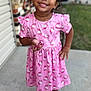 child, girl, smiling, pink_dress, unicorn_pattern, holding_candy, standing, porch, concrete, outdoor, house, footwear, crocs, earrings, curly_hair, happy, portrait, casual_clothing, daylight, background_blur