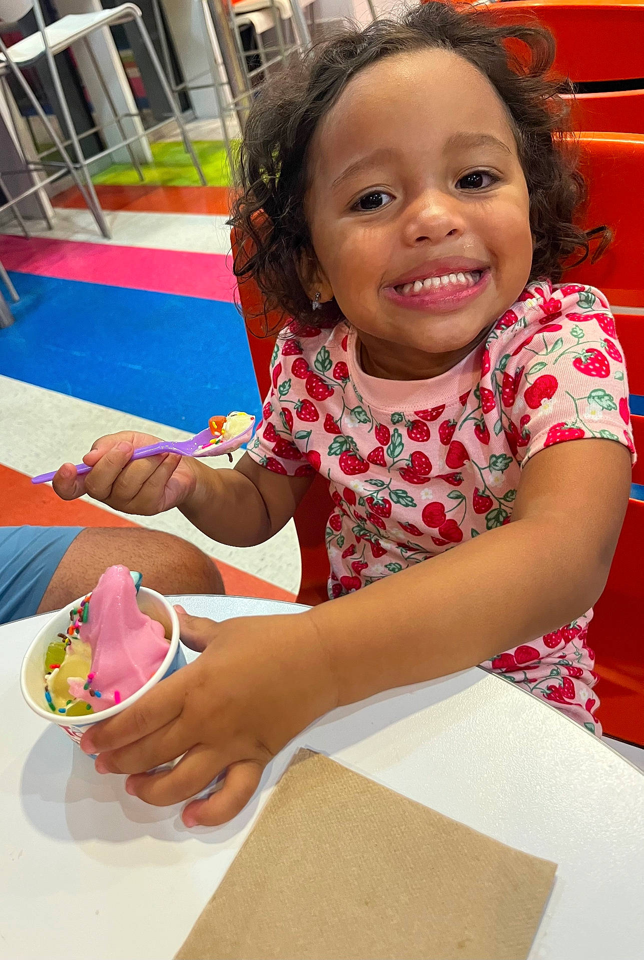 Zai'Lynn is registered to the contest to win money with this photo: casual_clothing, child, curly_hair, dessert, eating, food, frozen_yogurt, girl, happy, indoor, multicolored_floor, napkin, orange_chair, person, pink_shirt, smile, spoon, sprinkles, strawberry_pattern, table