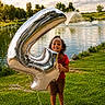 child, balloon, number_4, smiling, outdoor, grass, lake, water_fountain, trees, sunny, celebration, curly_hair, red_shirt, brown_shorts, sandals, happy, playful, nature, park, daytime