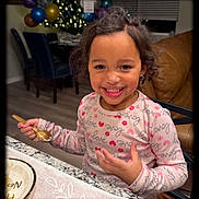 Zai'Lynn is registered to the contest to win money with this photo: child, smiling, curly_hair, spoon, table, plate, indoor, balloons, decorations, holiday, kitchen, happy, eating, person, clothing, long_sleeve, face, jewelry, earrings, home