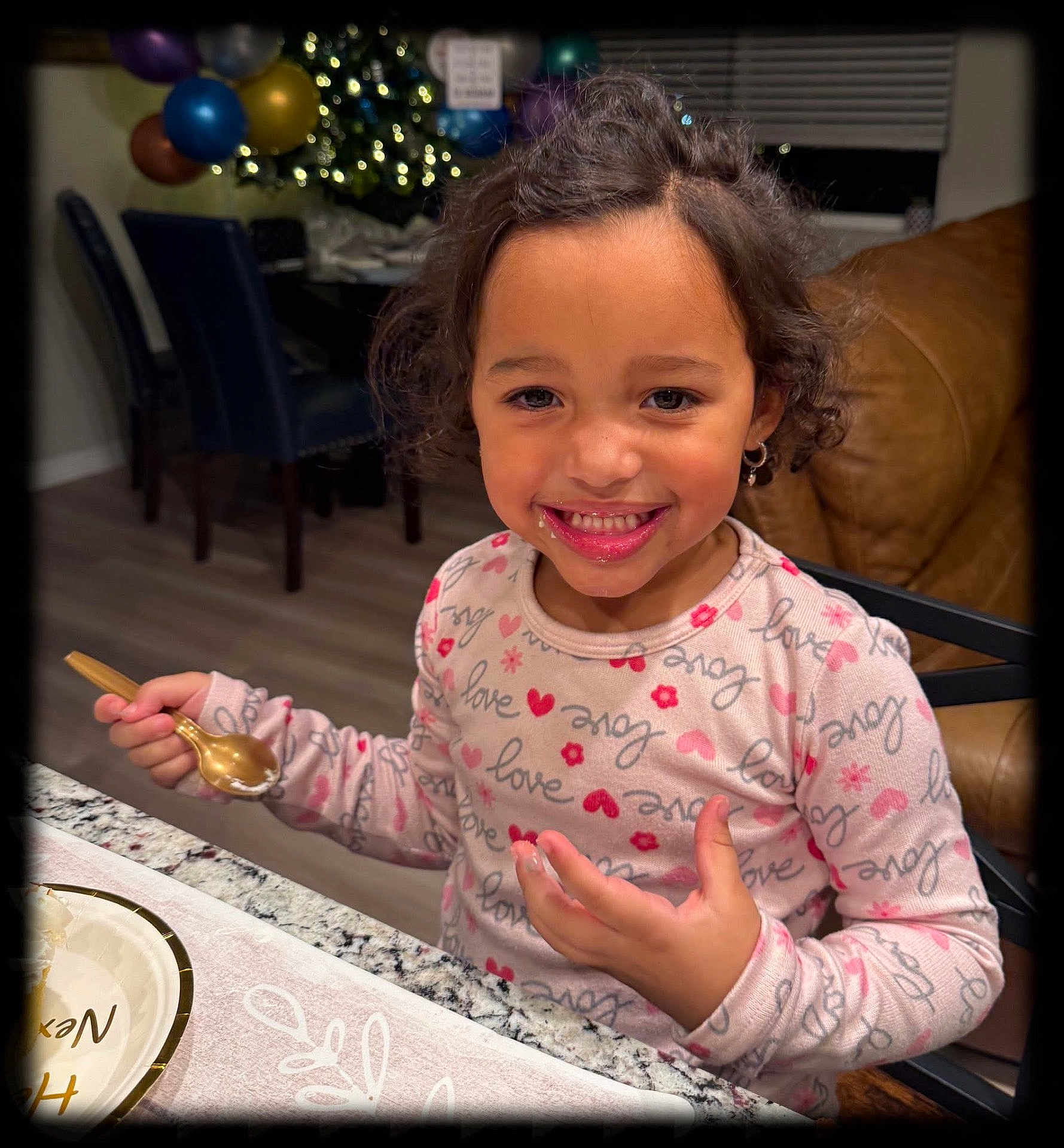 Zai'Lynn is registered to the contest to win money with this photo: child, smiling, curly_hair, spoon, table, plate, indoor, balloons, decorations, holiday, kitchen, happy, eating, person, clothing, long_sleeve, face, jewelry, earrings, home