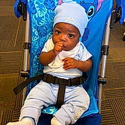 Xeiver is registered to the contest to win money with this photo: baby, child, infant, stroller, sitting, portrait, face, eyes, thumb_sucking, hat, beanie, white_clothing, blue, harness, carpet, indoor, smiling, baby_fashion, stitch, travel