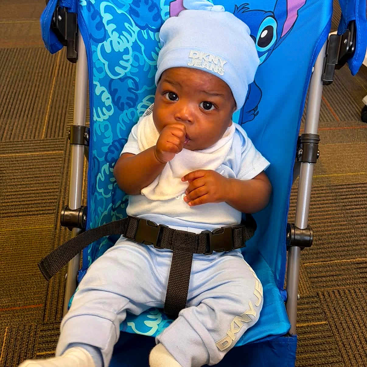 Xeiver is registered to the contest to win money with this photo: baby, child, infant, stroller, sitting, portrait, face, eyes, thumb_sucking, hat, beanie, white_clothing, blue, harness, carpet, indoor, smiling, baby_fashion, stitch, travel