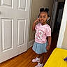 casual, child, clothing, denim_skirt, expression, flooring, footwear, girl, hair_accessories, hair_buns, hallway, indoor, person, pink_jersey, posing, sneakers, standing, white_door, wooden_floor, yellow_furniture