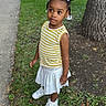 casual_clothing, child, curly_hair, cute, daylight, girl, grass, hair_bows, looking_away, nature, outdoor, portrait, sidewalk, standing, summer, tree, white_skirt, white_sneakers, yellow_striped_shirt, young
