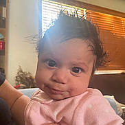 Adrianna is registered to the contest to win money with this photo: baby, child, infant, portrait, smiling, happy, pink_outfit, indoor, window, wooden_blinds, warm_lighting, close_up, face, eyes, curly_hair, sitting, adult_arm, home, cozy, photograph