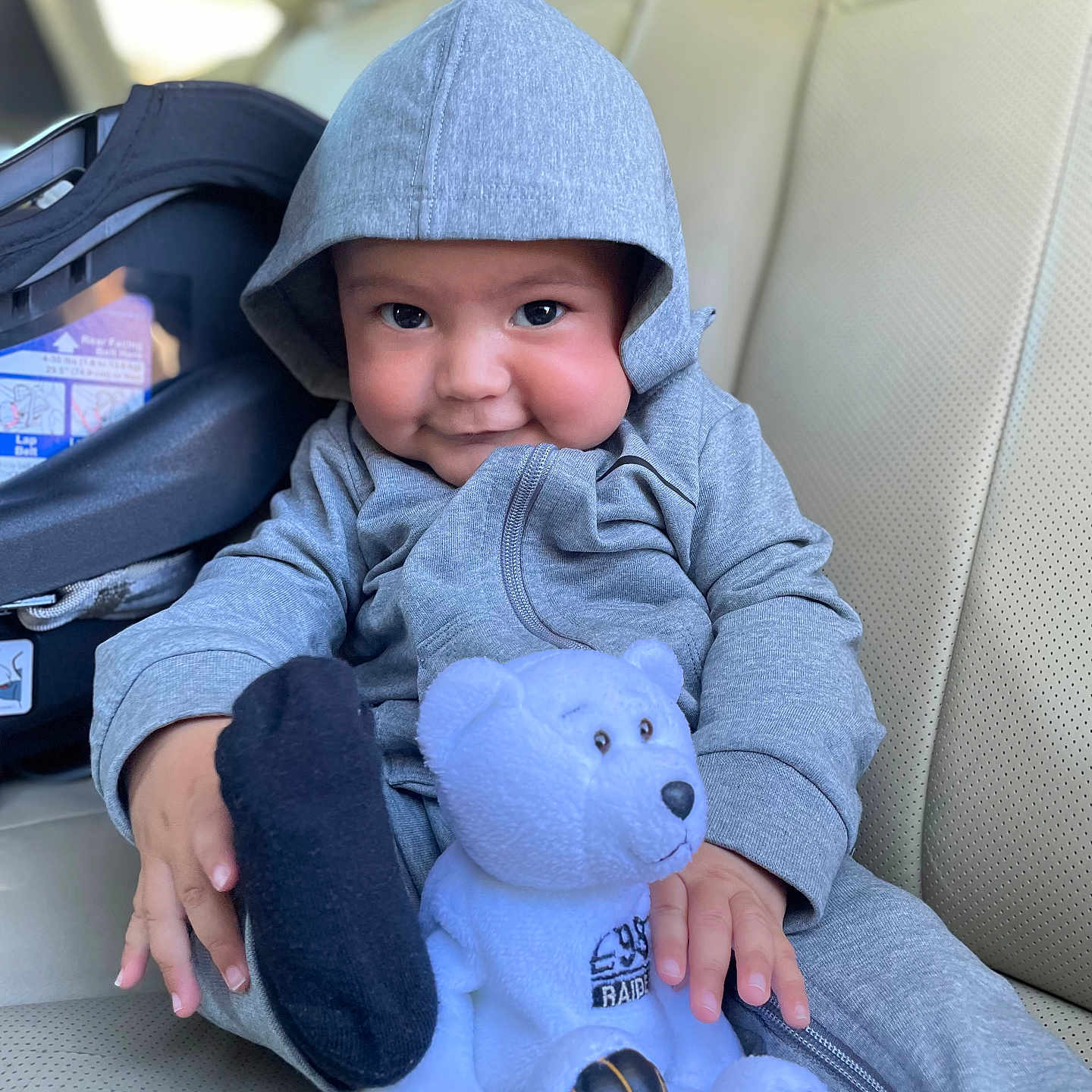 Kendrick is registered to the contest to win money with this photo: baby, beige_seat, car_interior, car_seat, child, clothing, cozy, cute, face, hand, hoodie, indoor, infant, plush, portrait, sitting, smile, teddy_bear, toy, young_child