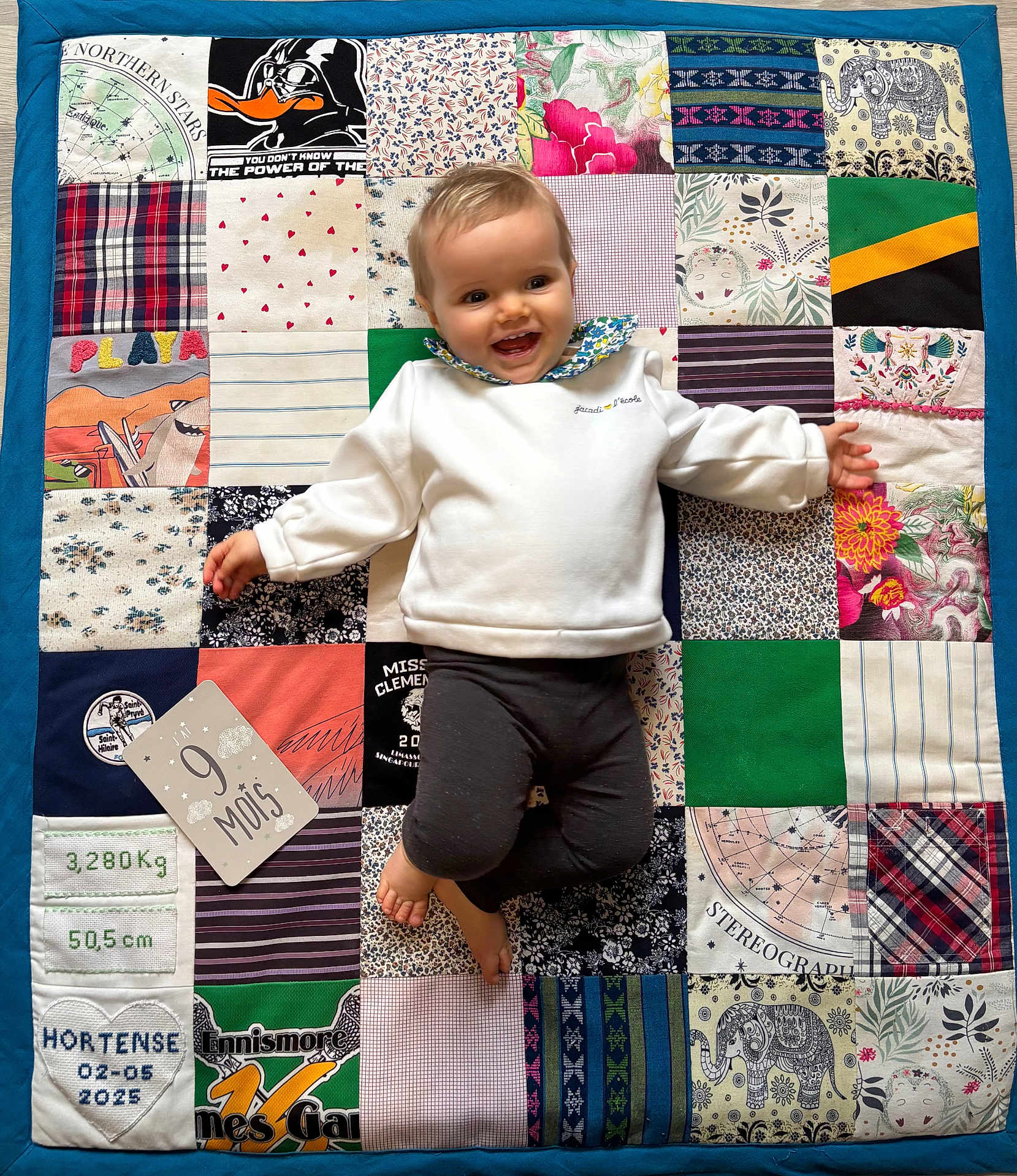 Hortense a rejoint le concours — aidez-le/la à gagner de superbes lots ! baby, child, smiling, patchwork_quilt, colorful, milestone_card, white_sweater, dark_pants, lying_down, happy, patterned_fabric, floral_pattern, striped_pattern, plaid_pattern, soft_texture, indoor, portrait, cute, infant, clothing