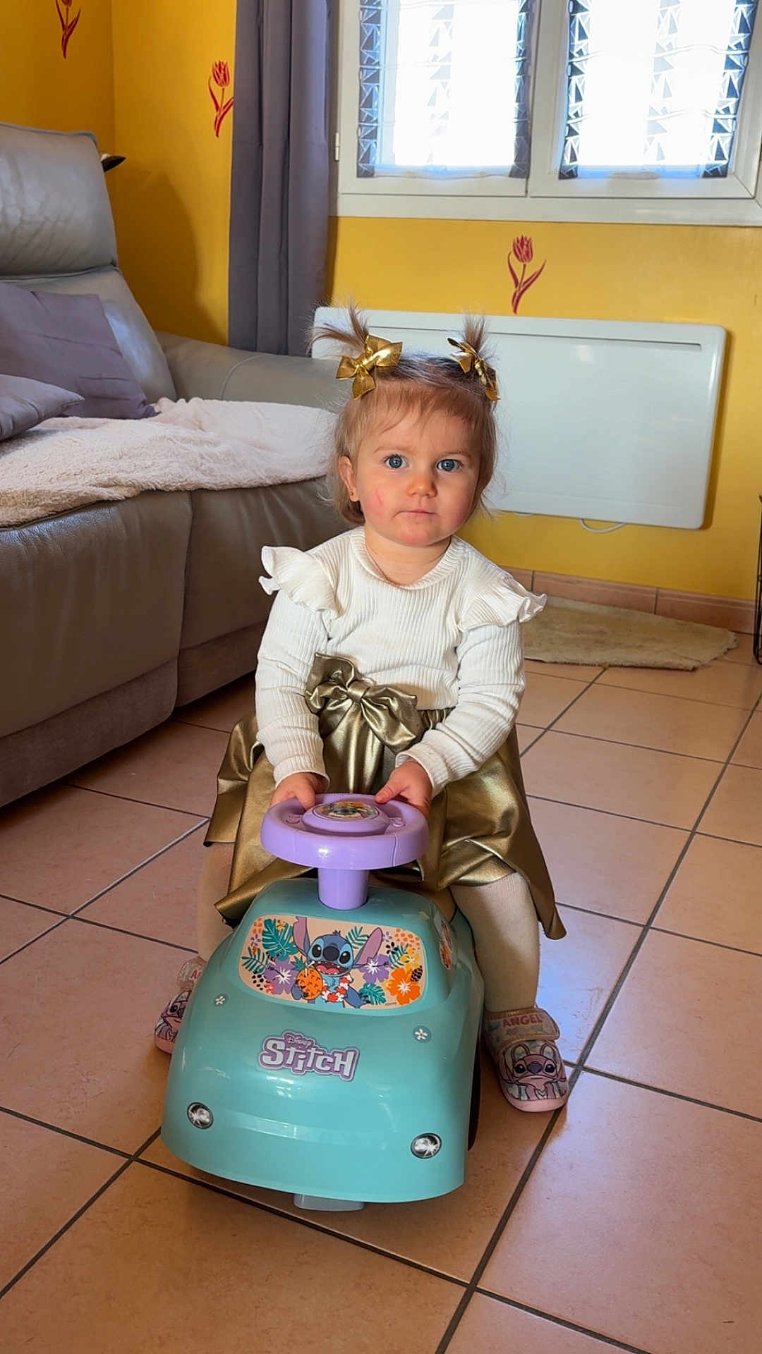 Mya a rejoint le concours — aidez-le/la à gagner de superbes lots ! toddler, child, indoor, toy, ride_on_toy, disney, stitch, blue, purple, golden_bow, white_sweater, gold_skirt, slippers, tile_floor, couch, blanket, window, curtain, home_interior, yellow_wall