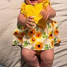 baby, child, smiling, yellow_dress, sunflower_pattern, infant, hands, legs, bed, blanket, happy, cute, person, portrait, indoor, clothing, skin, feet, comfort, playful