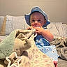 baby, child, hat, blue_dress, blanket, plush_elephant, toy, bed, pillow, marine_corps_flag, indoor, curious, infant, soft_toy, fabric, person, cute, sitting, nursery, cozy