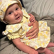 Emery joined the competition — help win amazing prizes! baby, child, infant, smiling, yellow_clothing, floral_pattern, bonnet, hand, blanket, bed, comfort, cute, portrait, skin, fingers, sitting, indoors, toddler, person, soft